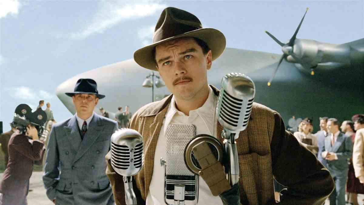 Un homme portant un chapeau marron avec une épaisse moustache (Leonardo DiCaprio) se tient devant une série de pieds de microphone avec un avion chromé géant visible derrière lui.