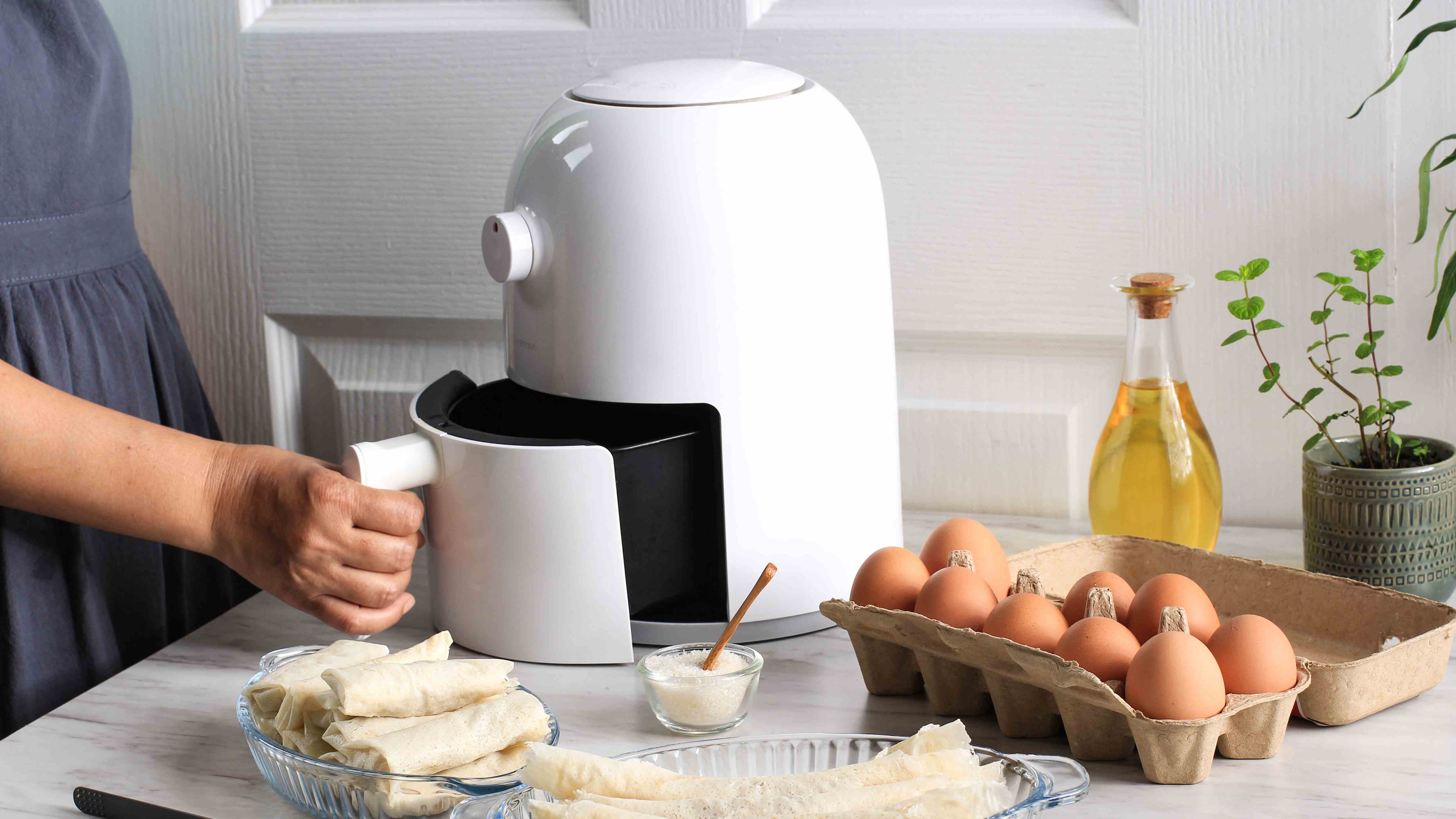 Une friteuse à air blanche avec le panier ouvert à la main sur une table avec des ingrédients environnants, y compris des œufs et de l'huile