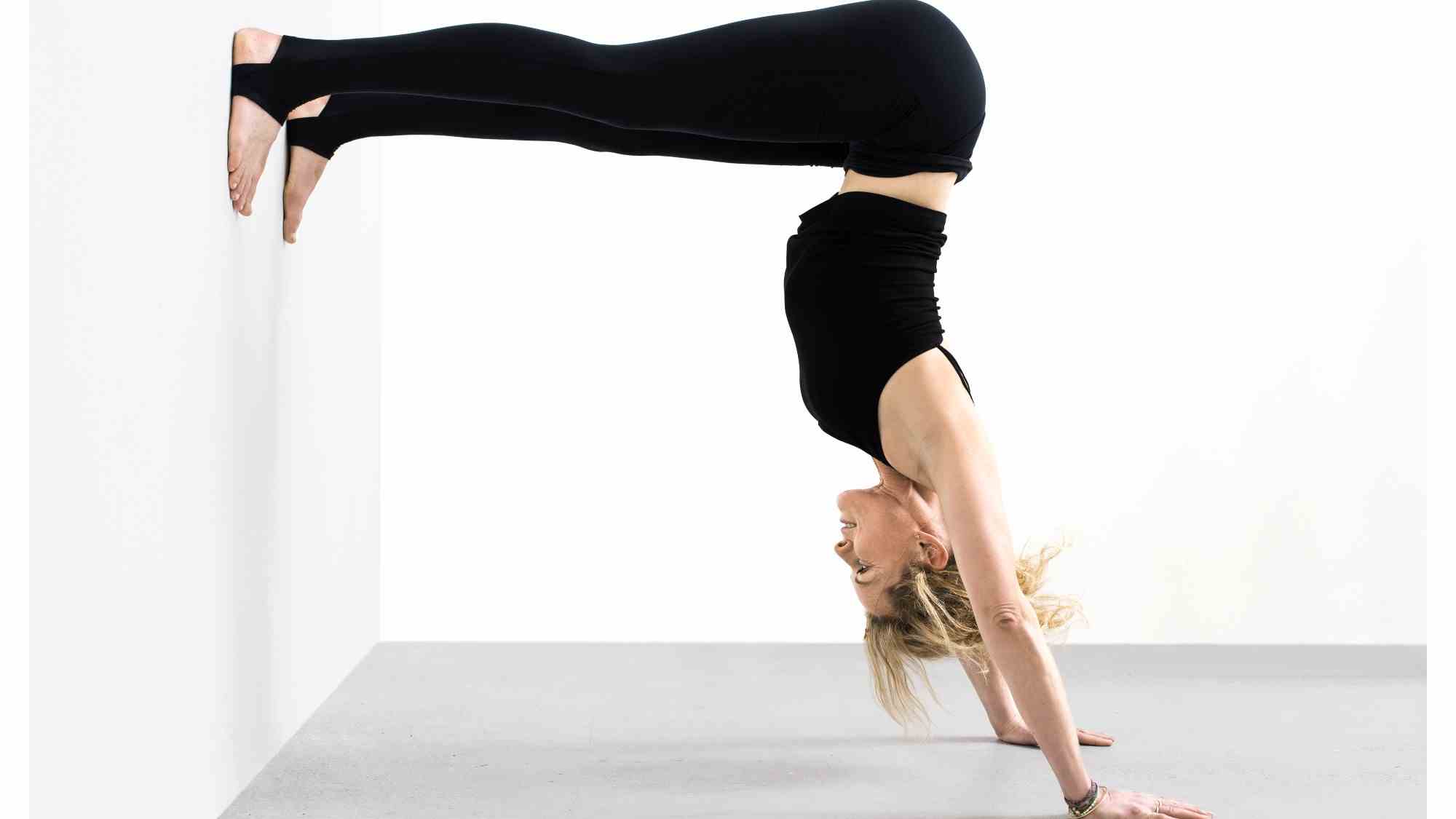 Femme face à un mur sur fond blanc effectuant un poirier avec les jambes droites contre le mur