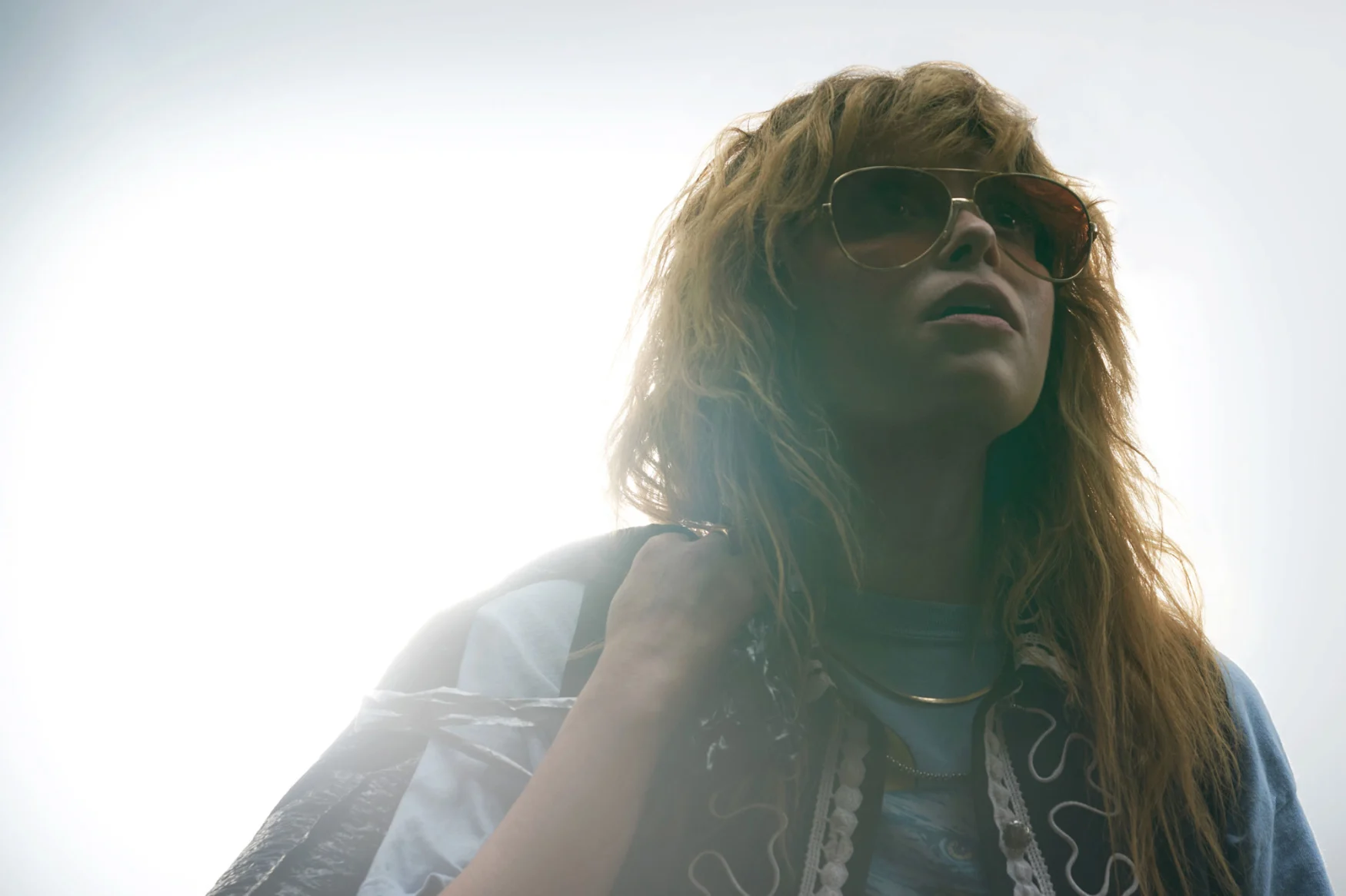 Image de Natasha Lyonne regardant vers le haut et vers la droite, hors caméra, dans une image surexposée si lumineuse qu'elle est la seule chose visible.  Elle porte des lunettes de soleil aviateur et tient sa veste par-dessus son épaule droite. 