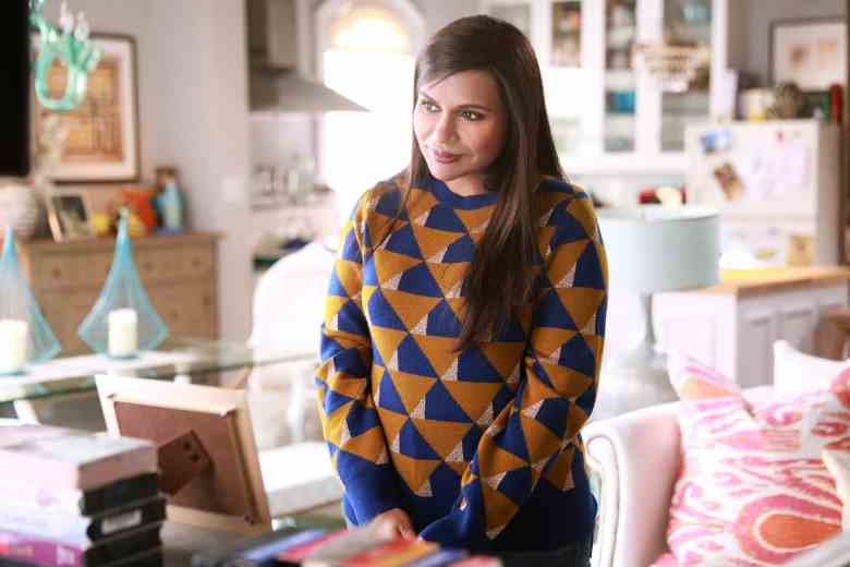 Une femme aux cheveux longs portant un pull à motifs marron et bleu marine, debout dans un bureau lumineux et coloré ;  encore de 