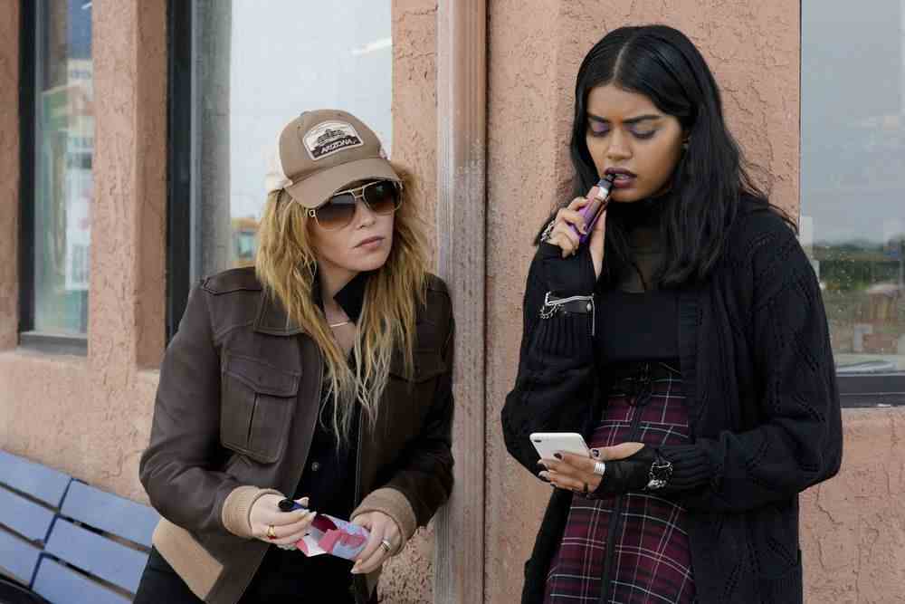 Natasha Lyonne dans le rôle de Charlie Cale, portant un chapeau de camionneur et de grosses lunettes de soleil, se penche pendant que Sara (Megan Suri) grignote sa vape et regarde son téléphone dans Poker Face