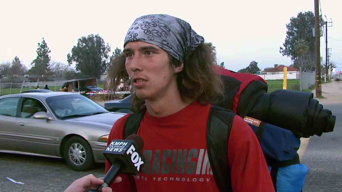 Un homme aux cheveux longs en chemise rouge portant un bandana gris sur la tête et un sac à dos parlant à un microphone dans un parking.