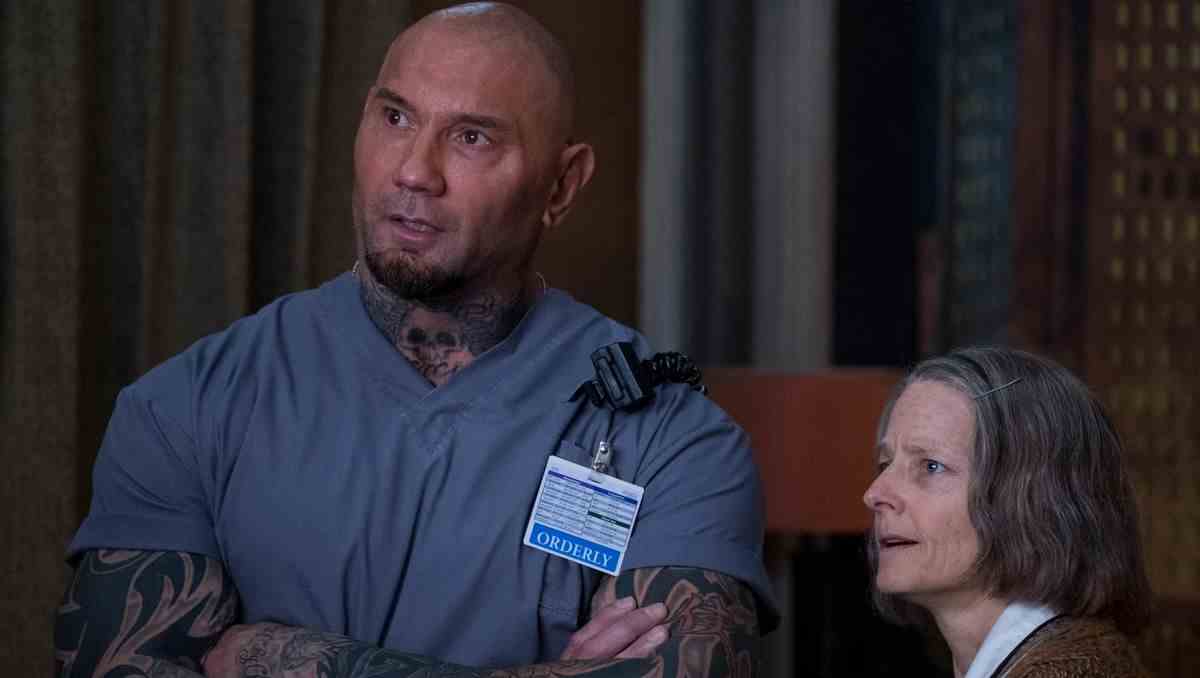 Dave Bautista croise les bras dans une blouse médicale avec une étiquette qui dit 