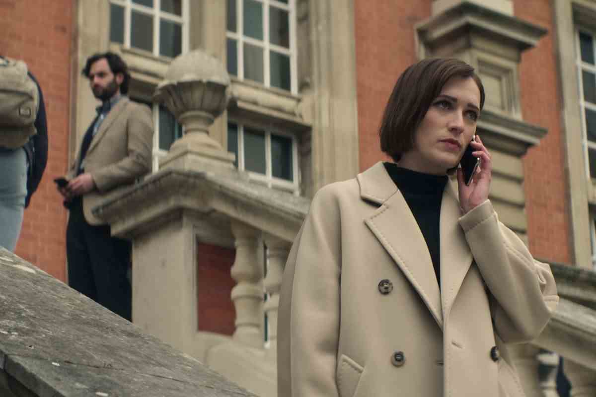 Joe se cache en arrière-plan en haut d'un escalier extérieur tandis que Kate se tient en bas au téléphone dans un trench-coat dans une scène de la saison 4 de Netflix's You.