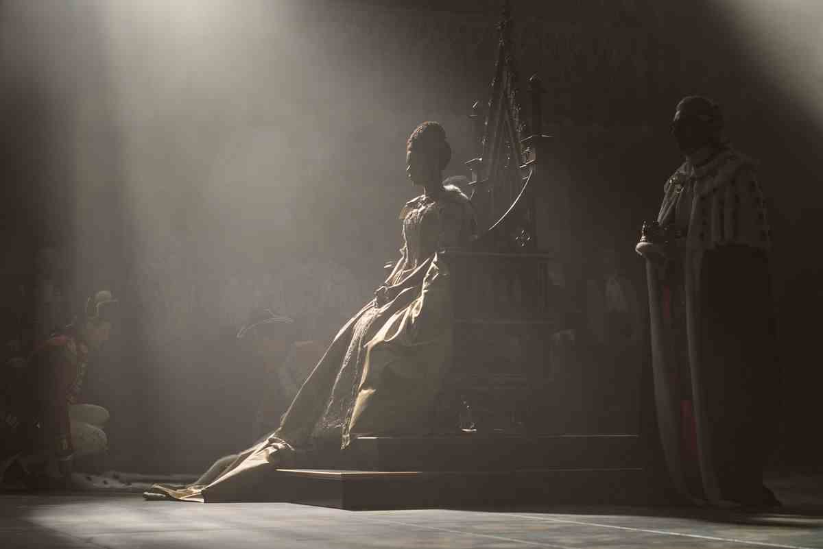 la reine charlotte assise sur son trône, des ombres dramatiques protégeant la plupart de ses traits, avec un rayon de soleil la frappant en plein centre