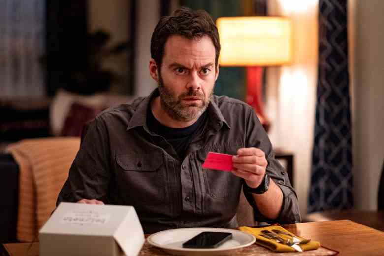 A man with dark hair and beard sits at a dining table, holding a red business card and looking horrified; still from "Barry."