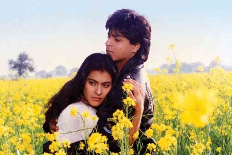 A man in a black leather jacket and a woman in a white Indian salwar-kameez hug in a field surrounded by yellow flowers; still of Kajol and Shah Rukh Khan in "Dilwale Dulhania Le Jayenge"