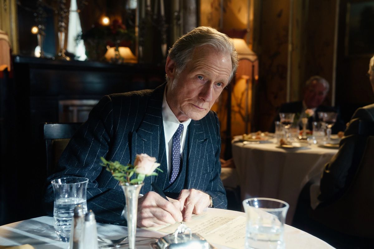 Bill Nighy, vêtu d'un costume à fines rayures, écrit quelque chose à la table d'un restaurant dans Living.