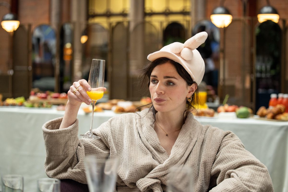 Aisling Bea boit ce qui ressemble à un mimosa près d'un buffet tout en portant quelque chose qui ressemble à un pyjama dans This Way Up.