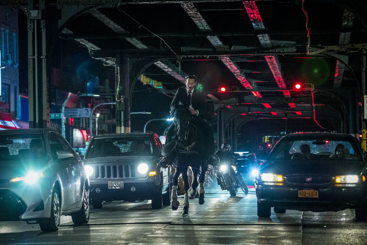 Keanu Reeves dans le rôle de John Wick, à cheval sous des voies ferrées surélevées la nuit, avec une moto le poursuivant dans la circulation