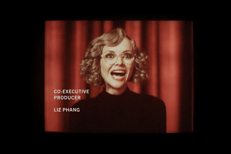 Medium shot of a woman with curly blonde hair and glasses laughing maniacally in front of a red curtain backdrop; still of Christina Ricci in "Yellowjackets"