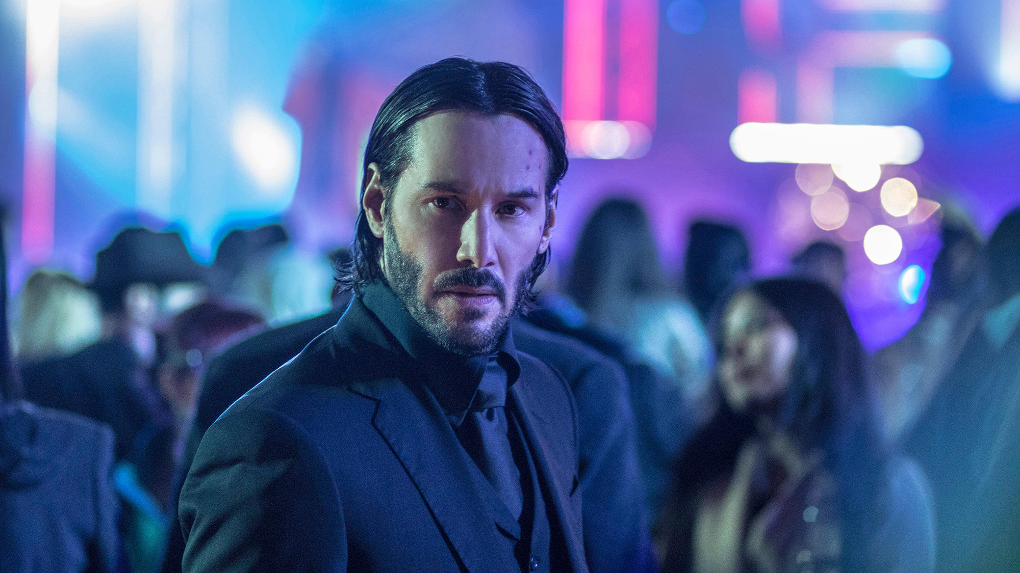 Keanu Reeves dans le rôle de John Wick, debout dans une foule de personnes avec un néon en arrière-plan dans John Wick: Chapitre 2