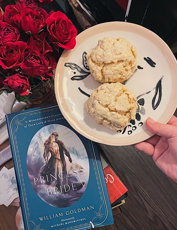 Une photo d'une main tenant une assiette avec deux scones dessus, tenue au-dessus de The Princess Bride et à côté de roses rouges