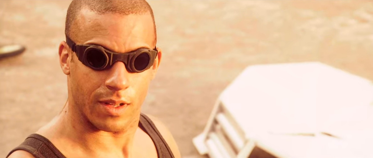 Vin Diesel dans le rôle de Richard B. Riddick face à la caméra portant des lunettes et un débardeur noir avec du sable en arrière-plan