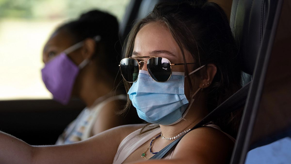 Une femme portant des lunettes de soleil et un masque bleu est assise au volant d'un véhicule avec un passager à côté d'eux portant un masque violet.