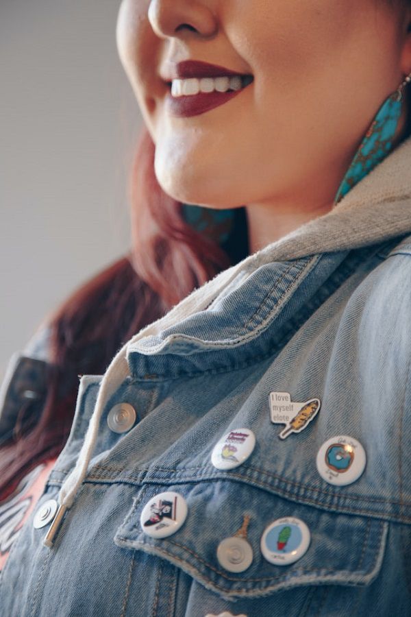 une femme souriante à la peau blanche portant du rouge à lèvres rouge foncé vue du nez vers le bas portant une veste ean lacée de diverses épingles et boutons