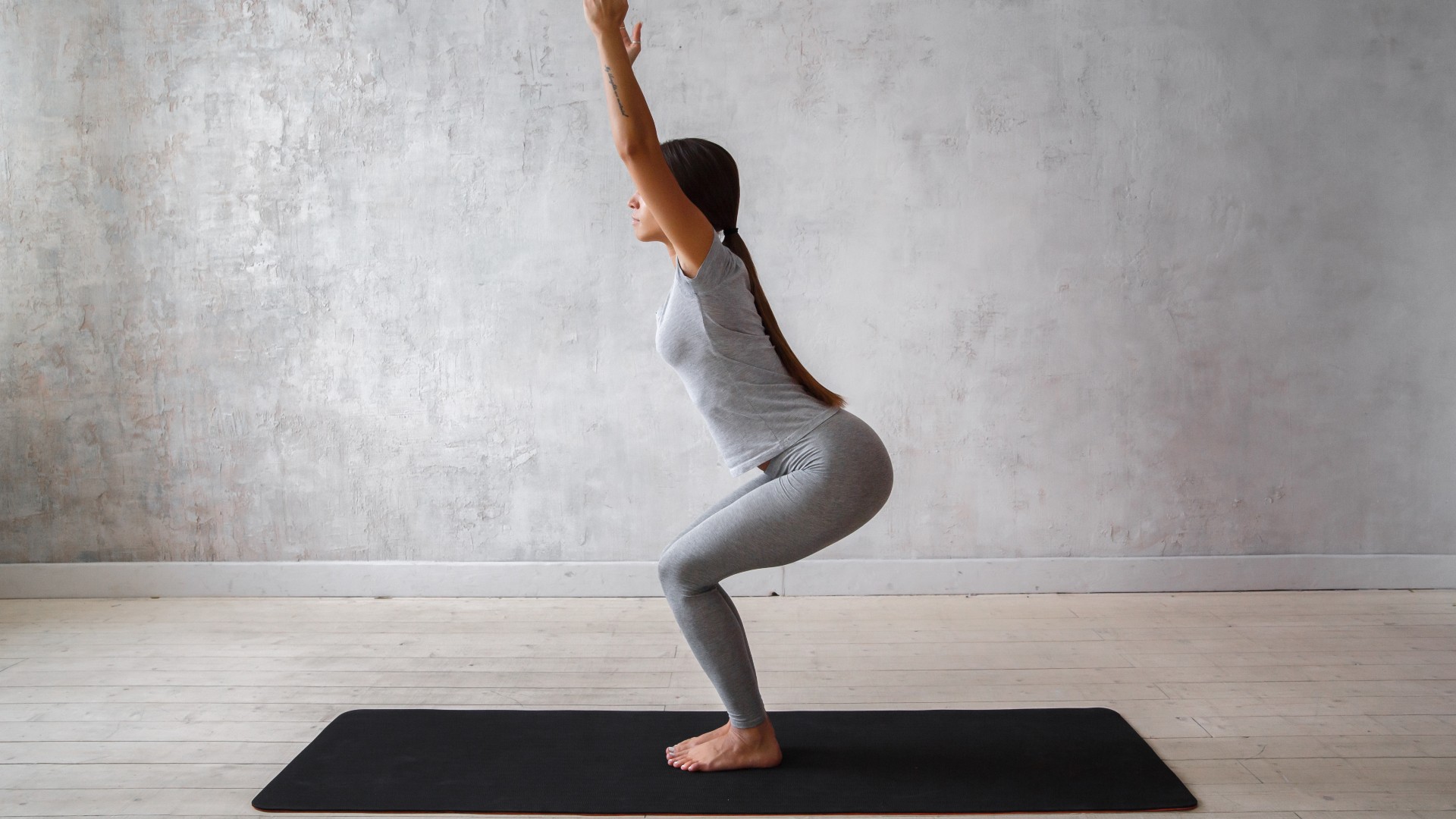 Femme sur son tapis de yoga sur fond gris avec les genoux pliés et les bras levés dans la pose de la chaise