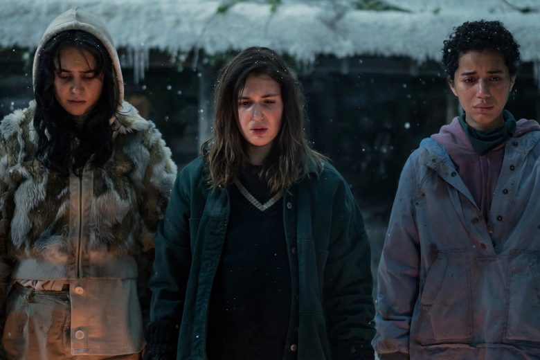 Three teen girls in winter outerwear standing outside a snowy cabin and looking down solemnly.