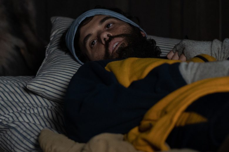 A man in yellow and blue winter athletic wear lays in bed with an empty expression; still from "Yellowjackets"