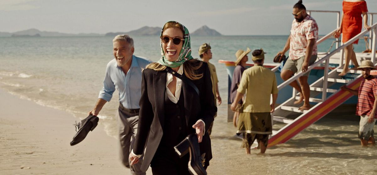 Un homme (George Clooney) et une femme portant un foulard sur la tête (Julia Roberts) rient en débarquant d'un bateau sur une plage et tenant leurs chaussures à la main.