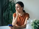 Triste et frustrée, une jeune femme brune pleure avec un smartphone à la main alors qu'elle est assise sur la chaise de l'appartement