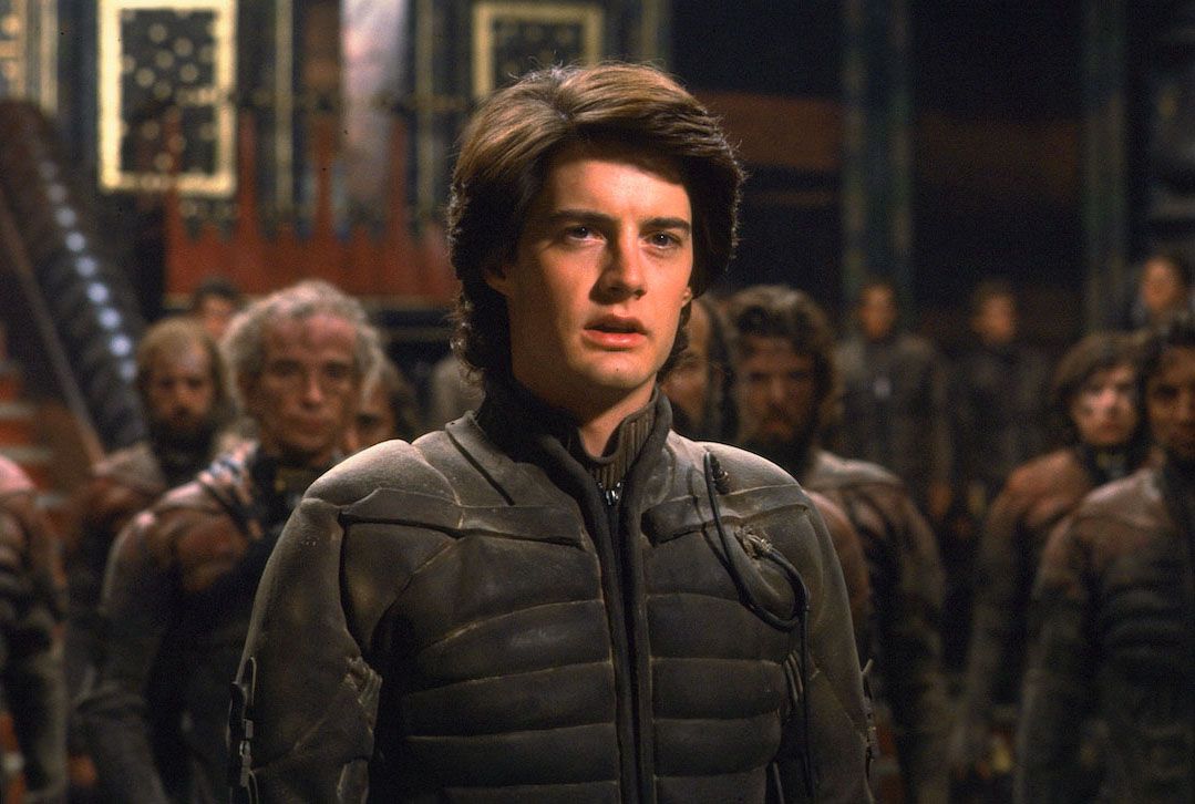 Un jeune homme ( Kyle Maclachlan ) aux cheveux bruns vêtu d'un costume high-tech poussiéreux se tient dans un atrium entouré de personnes vêtues de costumes similaires dans Dune (1984).