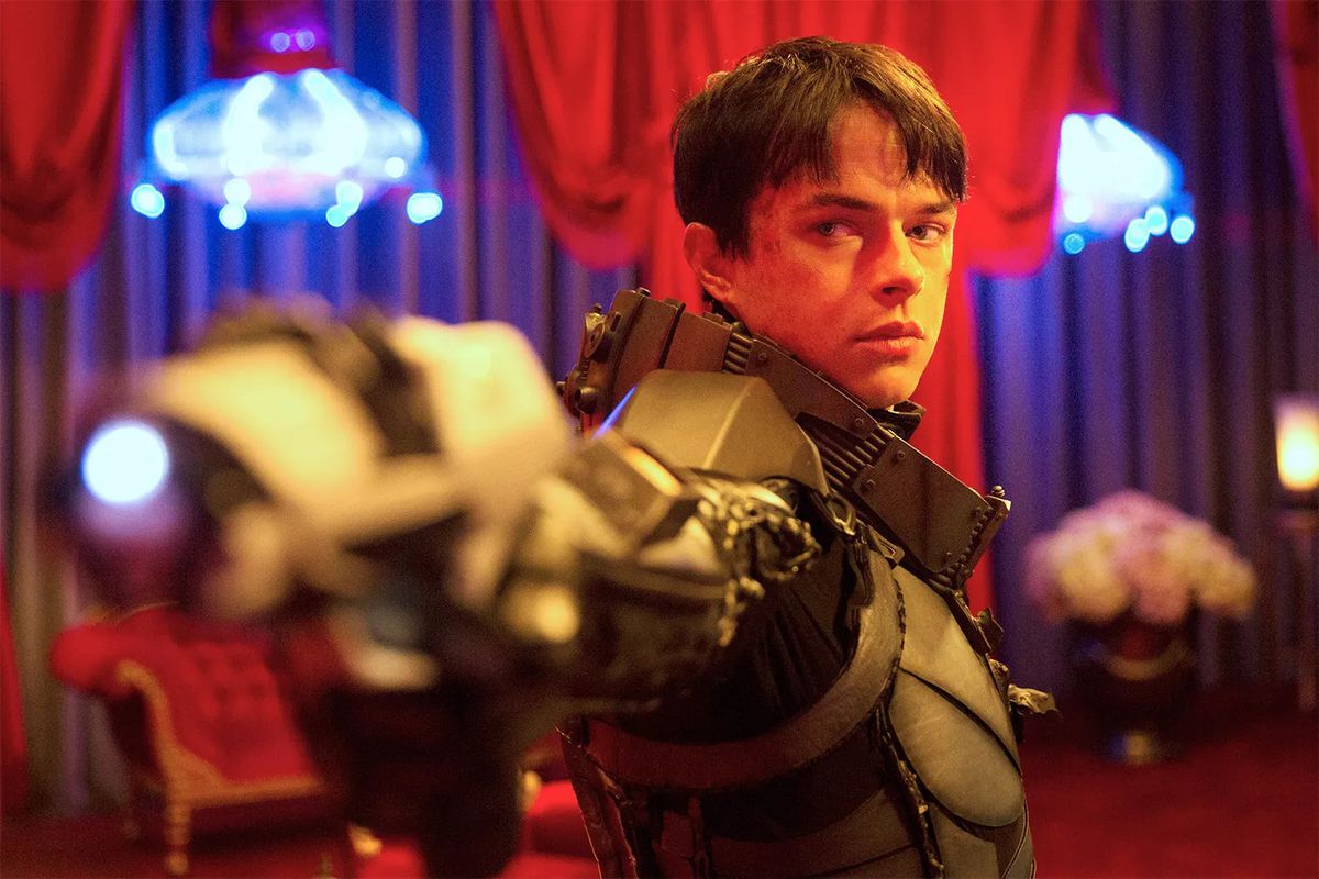 Un jeune homme (Dane DeHaan) aux cheveux noirs courts dans une combinaison spatiale high-tech pointe un pistolet lumineux dans une pièce éclairée en rouge avec des lustres bleus en arrière-plan dans Valerian and the City of a Thousand Planets.