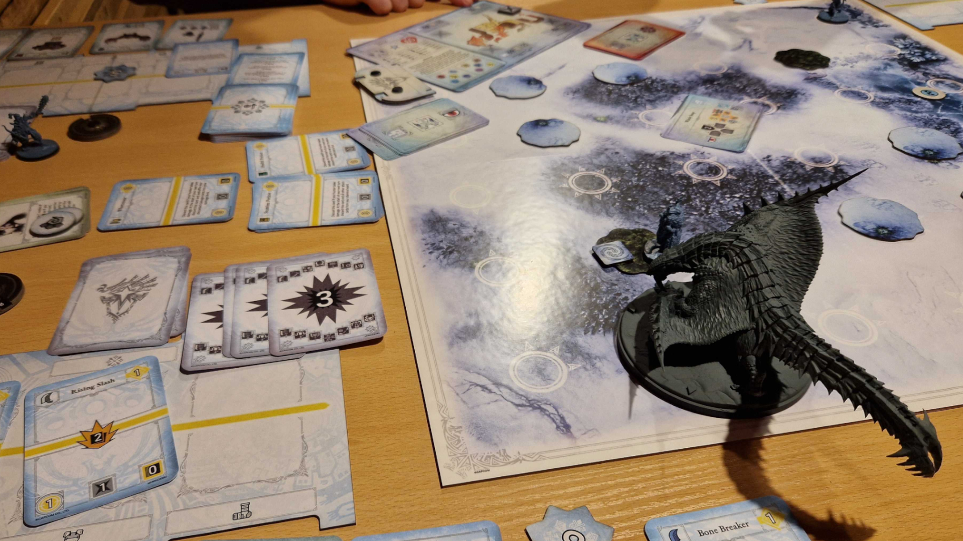 Un aperçu du Monster Hunter World Iceborne : le plateau de jeu de société, des jetons, des miniatures et des cartes sur une table en bois