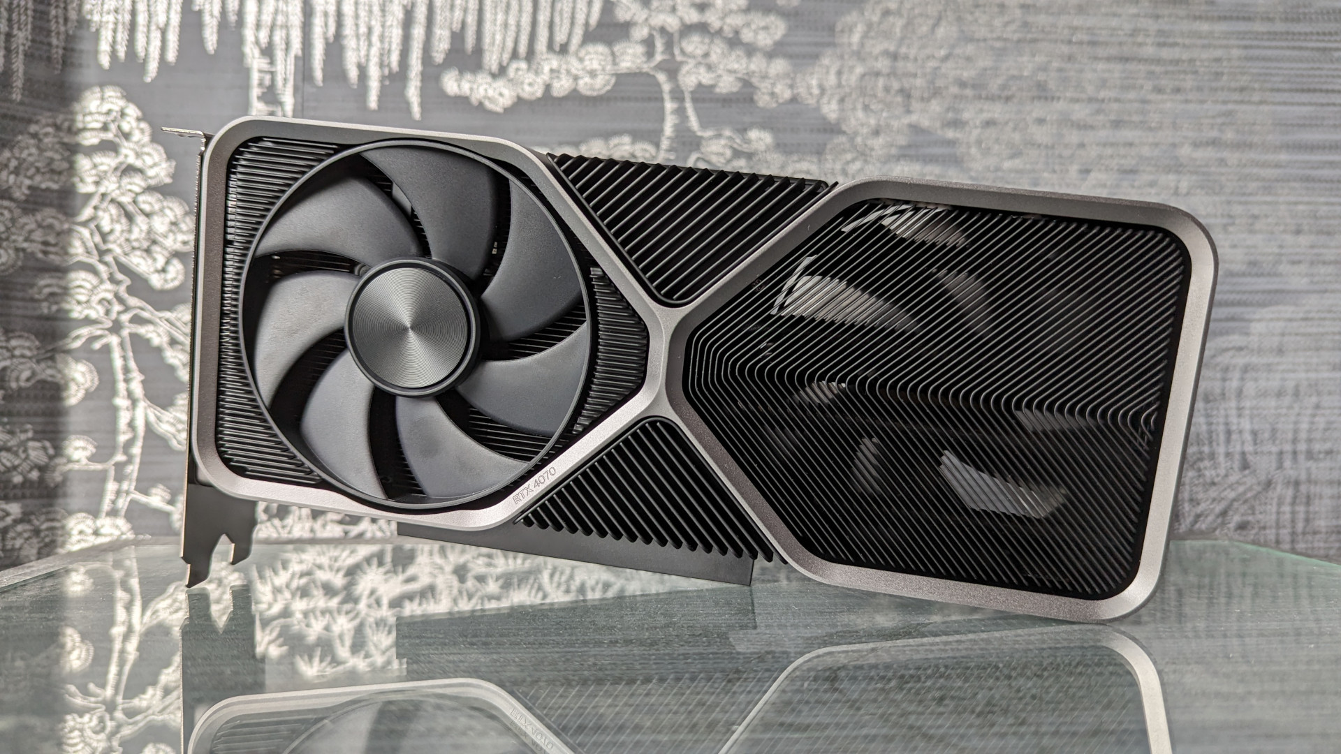 Test Nvidia GeForce RTX 4070 : Une carte graphique Founders Edition se reflétant sur une surface en verre, sur un fond gris-argenté