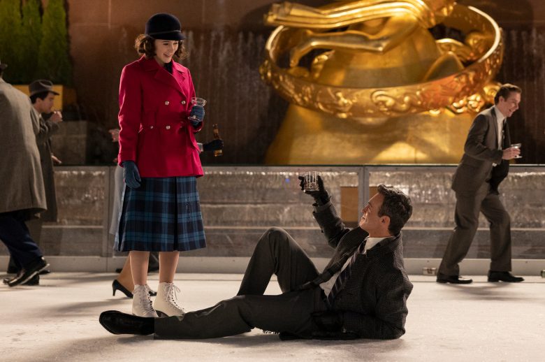 A man laying on an ice rink lifts his whiskey glass up at a woman in a plaid blue dress and magenta coat who stands looking down at him; still from "The Marvelous Mrs. Maisel"