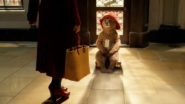 Paddington Bear sits on a suitcase; Mrs. Brown's legs appear in the foreground of the shot