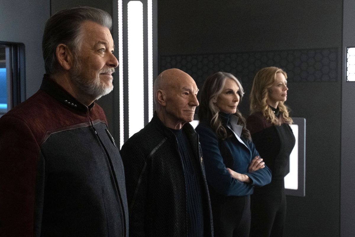 Ltr: Jonathan Frakes comme Will Riker, Patrick Stewart comme Jean Luc Picard, Jeri Ryan comme Seven of Nine et Gates McFadden comme Dr Beverly Crusher dans la saison 3 de Star Trek: Picard.  Ils se tiennent côte à côte en souriant.  Riker et Seven sont en uniforme.