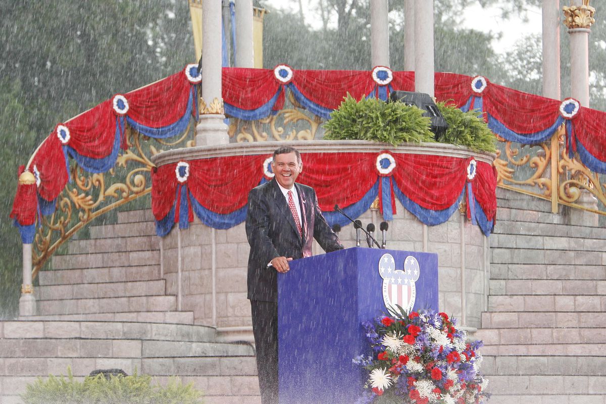 Le sénateur américain Mel Martinez se tient souriant sous une pluie battante sur un podium extérieur avec une tête de Mickey Mouse avec le drapeau américain dessus lors d'une cérémonie de naturalisation de masse à Walt Disney World en Floride le 4 juillet 2007