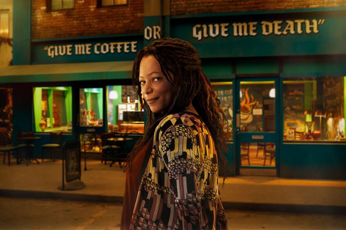 une femme avec des dreadlocks sombres sourit devant un magasin qui dit DONNEZ-MOI DU CAFÉ OU DONNEZ-MOI LA MORT