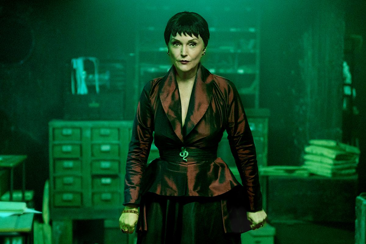 Miranda Richardson porte un combo blazer rouge bien ajusté et jupe crayon noire, avec ses cheveux dans une coupe au bol noire agitée.  Elle se tient dans un bureau sombre éclairé en vert. 