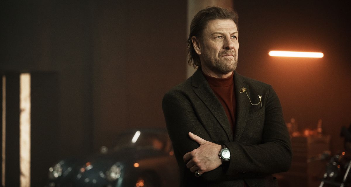 Alman Kido (Sean Bean), un homme pensif en costume, se tient dans un garage faiblement éclairé plein de voitures brillantes et chères et regarde vaguement hors de l'écran dans Knights of the Zodiac