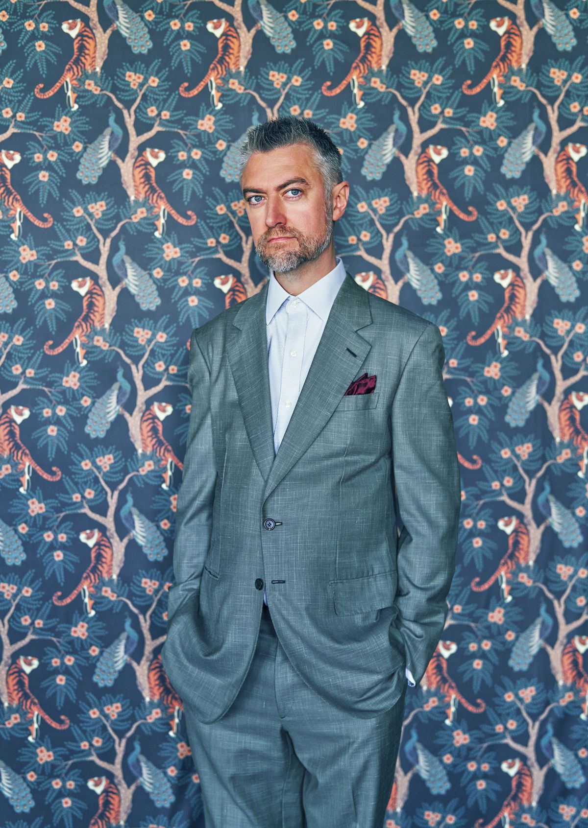 L'acteur Sean Gunn en costume bleu, debout sur un arrière-plan animé avec un motif répété de paons dans les arbres avec des tigres en dessous