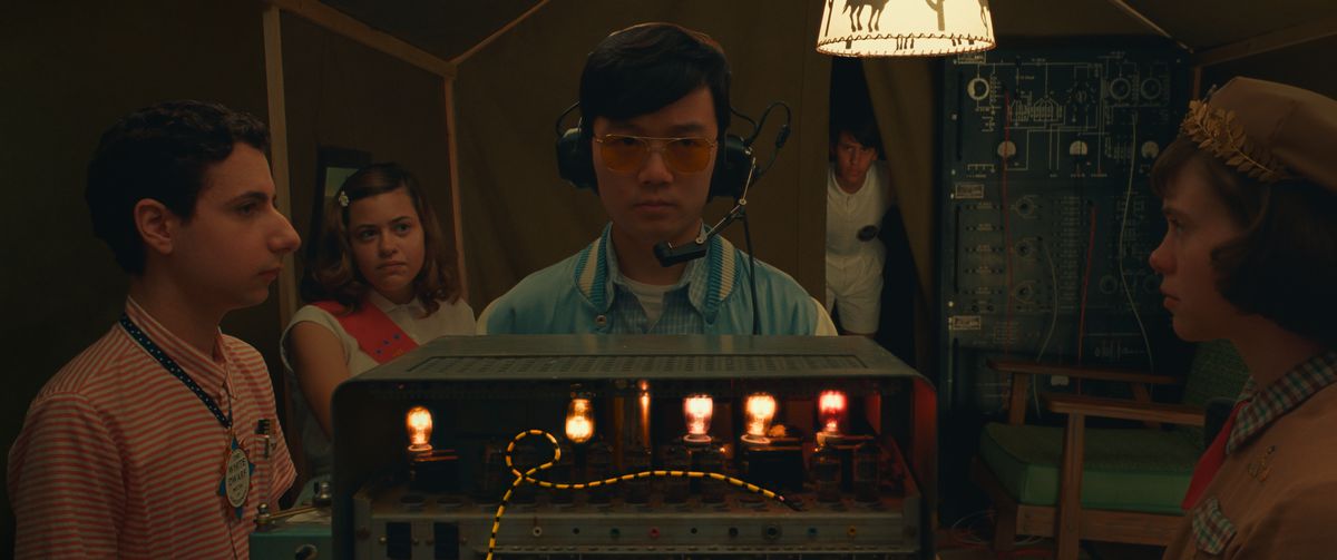 Dans une pièce très sombre, un groupe de jeunes (Jake Ryan, Grace Edwards, Ethan Josh Lee, Aristou Meehan et Sophia Lillis) se tient autour d'une radio ancienne couverte de lumières et de cadrans dans Asteroid City de Wes Anderson.