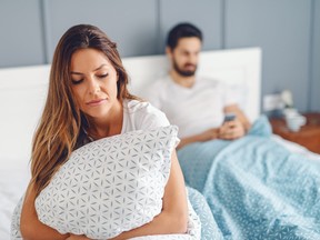 Femme insatisfaite tenant un oreiller et assise sur le lit pendant que son mari est sur un smartphone.