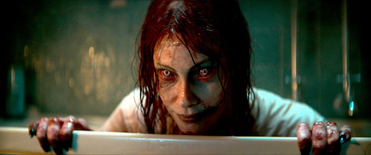 Ellie (Alyssa Sutherland), possédée et transformée en Deadite aux yeux rouges et jaunes, aux cheveux gras et au visage crasseux, sourit étrangement par-dessus une barrière dans Evil Dead Rise