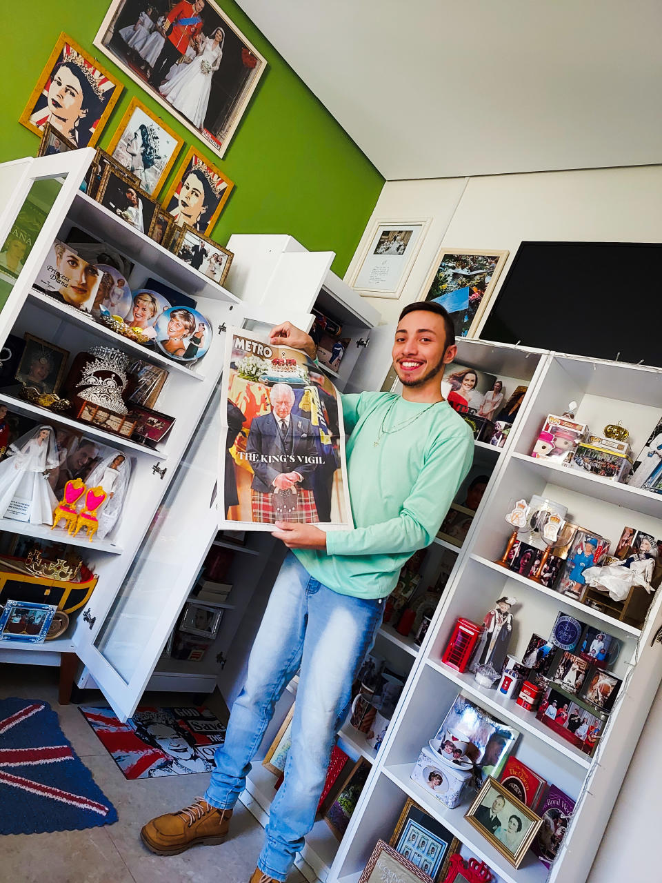 Savio Valei debout devant ses étagères pleines de souvenirs royaux tenant un journal avec une photo du roi Charles III en première page