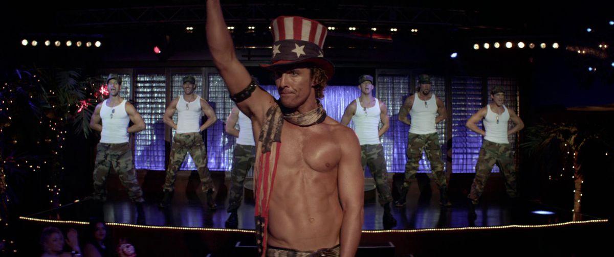 Un Matthew McConaughey torse nu avec un chapeau à rayures rouges, blanches et bleues avec des étoiles et un mouchoir se tient devant une scène d'hommes en chemises blanches et pantalons à motif camouflage au garde-à-vous dans Magic Mike.