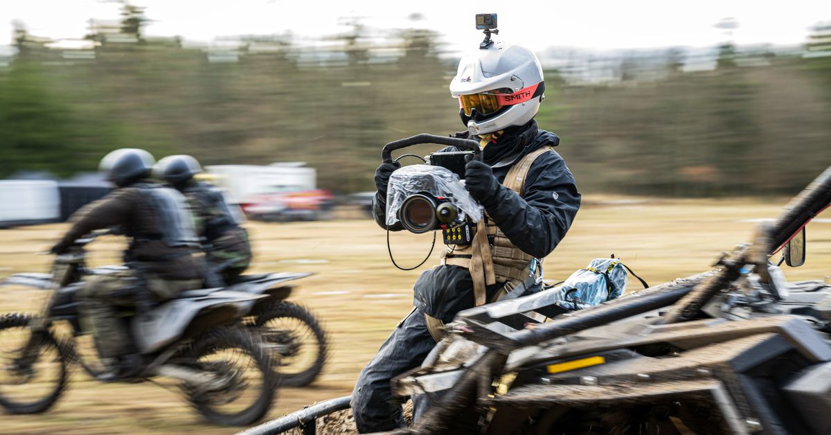 Le réalisateur Sam Hargrave, dans un casque de moto, des lunettes de protection et un harnais de sécurité, tenant.  une caméra et portant une seconde sur sa tête, s'assoit à l'avant d'un véhicule en mouvement pendant le tournage d'une séquence dans Extraction 2