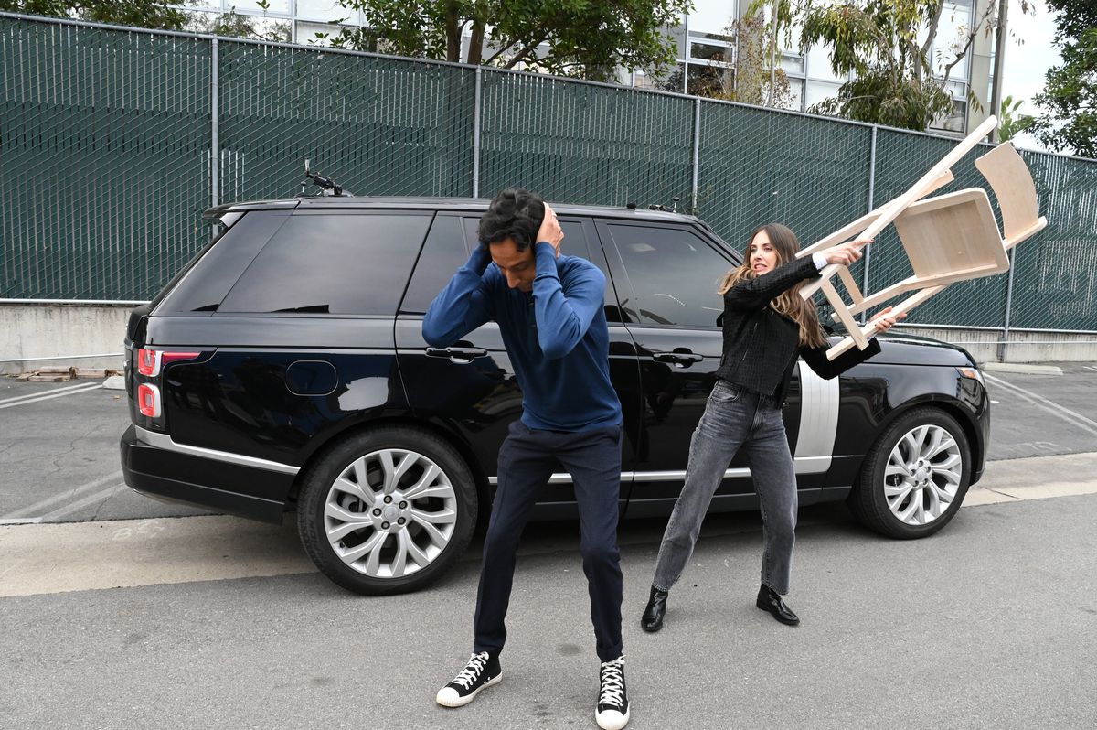 Danny Pudi se tient les oreilles et s'accroupit un peu tandis qu'Alison Brie arrive derrière lui avec une chaise sur le point de le frapper.  La voiture Carpool Karaoke est derrière eux. 