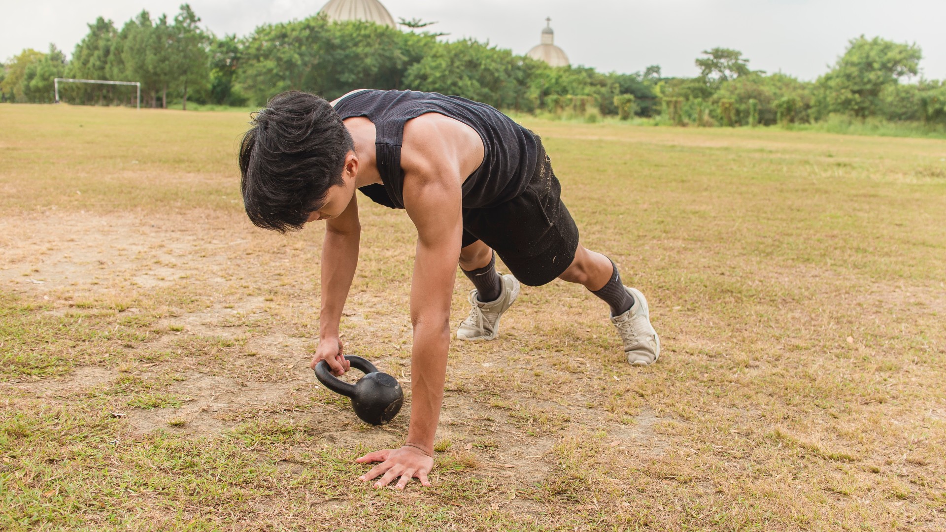 Homme sur l'herbe en position de planche déplaçant un kettlebell d'un côté à l'autre