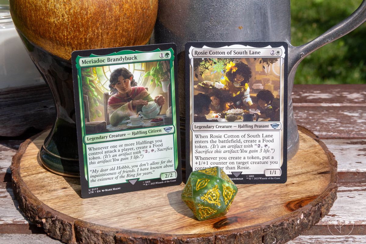 Une image montrant deux cartes Magic: the Gathering côte à côte avec un dé.  Les cartes illustrées sont Meriadoc Brandybuck et Rosie Cotton de South Lane de l'ensemble Tales of Middle-earth.