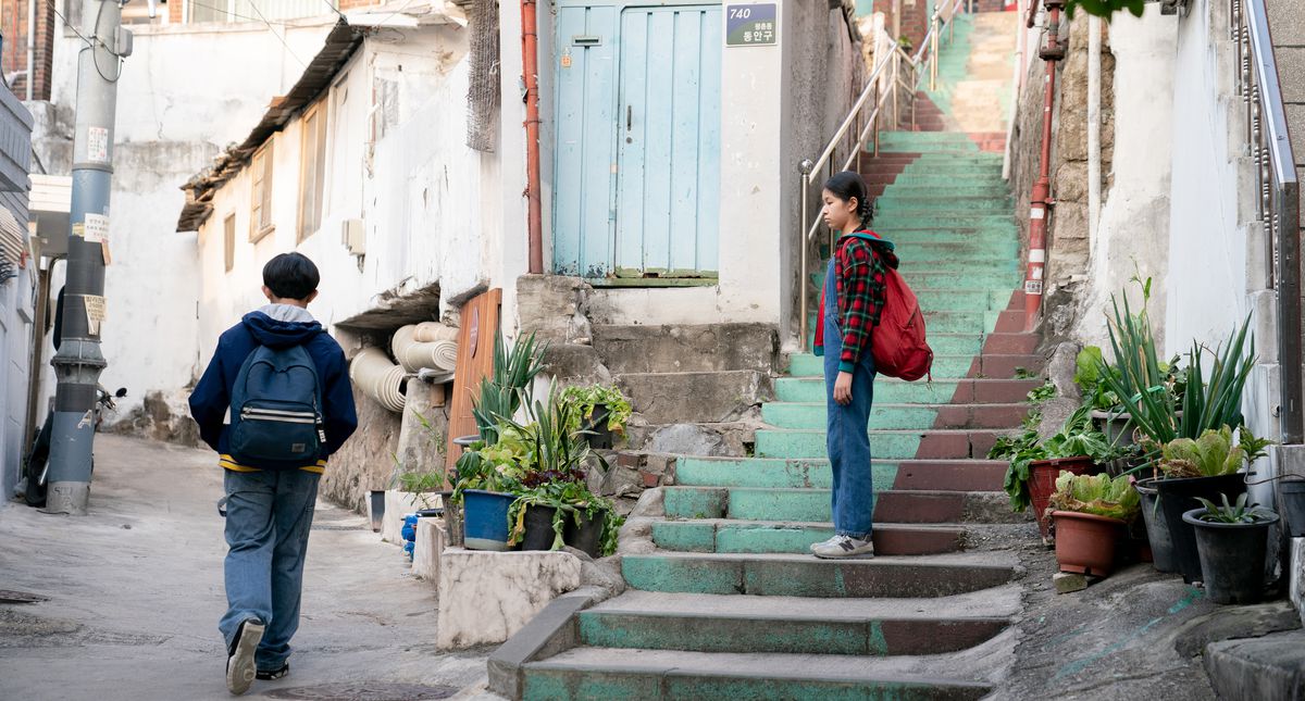 Deux enfants rentrent de l'école à pied.  La fille attend dans l'escalier, regardant le garçon s'éloigner.