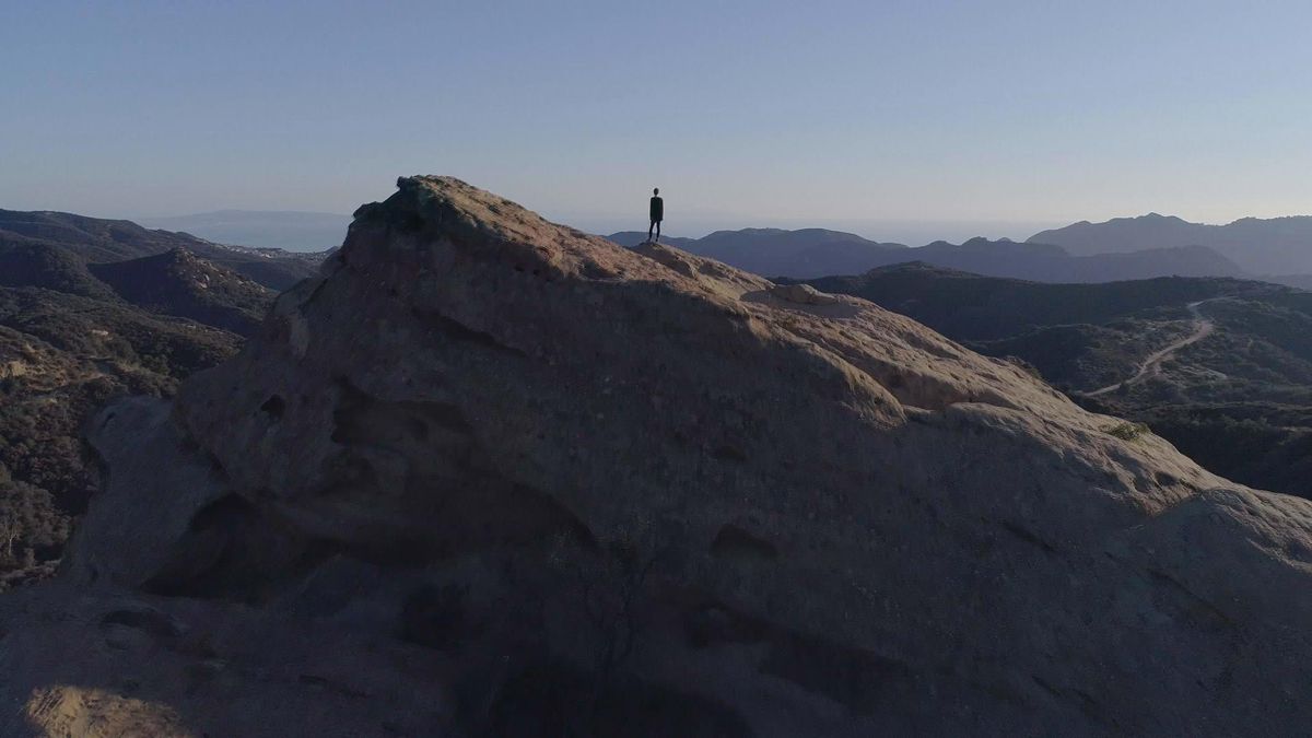 Narcissa Wright en silhouette minuscule, debout sur un vaste rocher incliné surplombant des montagnes basses dans une photo de Break the Game