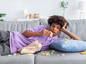 Adolescent ennuyé allongé sur un canapé avec des collations dispersées, mangeant des frites, regardant une émission ou un film ennuyeux à la télévision à la maison.